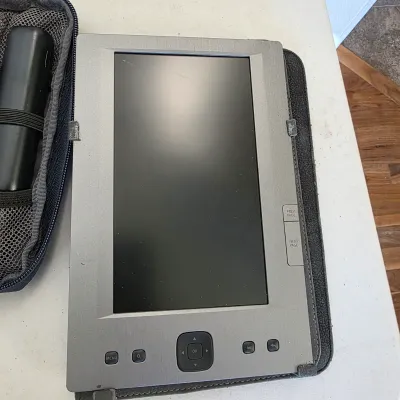 Sale Photo Thumbnail #158: Vintage silver-tone e-reader in a black protective folio case, featuring physical navigation buttons and page-turn controls.