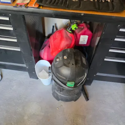 Sale Photo Thumbnail #152: Black wet/dry shop vacuum stored beneath a workbench alongside plastic gas cans and a white bucket, showing light wear.