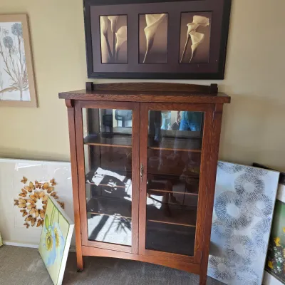 Sale Photo Thumbnail #96: Antique Arts and Crafts style oak curio cabinet featuring glass-paneled double doors and interior shelving, displayed alongside framed wall art.