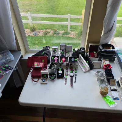 Sale Photo Thumbnail #162: Collection of men's and women's wristwatches, jewelry boxes, small purses, and accessories arranged on a white folding table near a window.