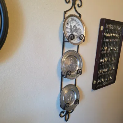 Sale Photo Thumbnail #69: Black wrought iron wall-mounted plate rack holding three decorative collector plates, mounted on the wall alongside a framed souvenir spoon display case.