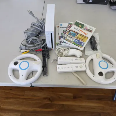 Sale Photo Thumbnail #112: Nintendo Wii gaming console bundle including the white console, sensor bar, power supply, AV cables, two Wii remotes with wrist straps, a Nunchuk attachment, two steering wheel accessories, and boxed Wii games.
