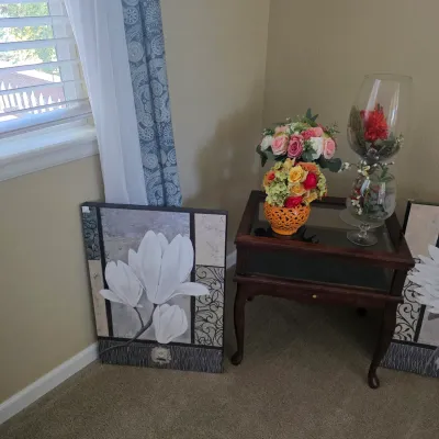 Sale Photo Thumbnail #79: Traditional wooden end table featuring glass accent decor and artificial floral arrangements, displayed alongside two modern canvas wall art pieces with white floral motifs.