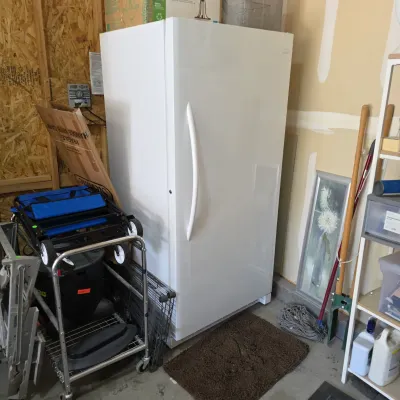 Sale Photo Thumbnail #3: White upright freezer stored in a garage, alongside a blue folding utility wagon and household storage items.