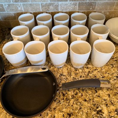 Sale Photo Thumbnail #186: Large set of modern white ceramic coffee mugs arranged alongside nonstick frying pans on a granite countertop.