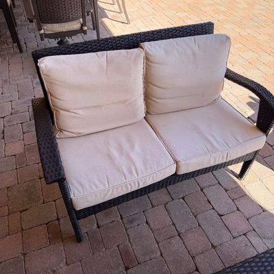 Sale Photo Thumbnail #246: Outdoor dark brown resin wicker loveseat with beige seat and back cushions, set on a brick paved patio.