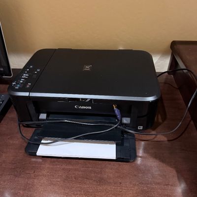 Sale Photo Thumbnail #140: Black Canon Pixma all-in-one inkjet printer with power cord, resting on a wooden desk surface.