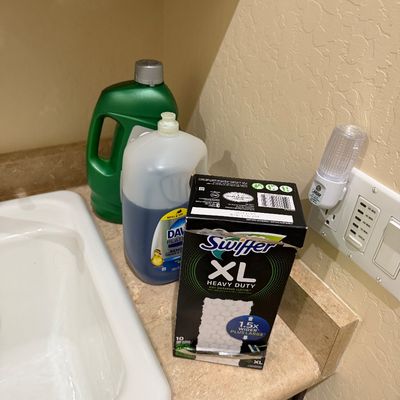 Sale Photo Thumbnail #109: Assortment of household cleaning supplies, including a box of Swiffer XL Heavy Duty dry sweeping cloths, Dawn Platinum dish soap, and laundry detergent next to a sink.