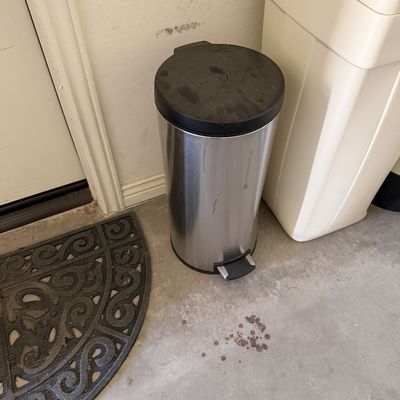 Sale Photo Thumbnail #92: Cylindrical stainless steel step trash can with a black plastic lid, showing minor wear and surface marks.