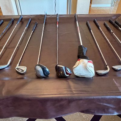 Sale Photo Thumbnail #253: Assorted collection of modern golf clubs laid out on a table, including irons, woods, and a TaylorMade Burner driver with a headcover.