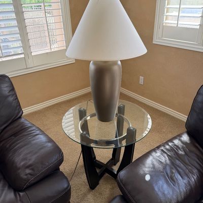 Sale Photo Thumbnail #238: Contemporary round glass-top end table with a dark wooden X-base, featuring a modern champagne-colored ceramic table lamp with a white shade, flanked by two dark brown leather armchairs.