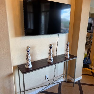 Sale Photo Thumbnail #35: Modern narrow console table with a dark wood top and slim metal base, displayed under a wall-mounted flat-screen television with three decorative ceramic tequila bottles on top.