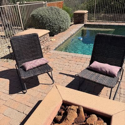 Sale Photo Thumbnail #241: Pair of dark brown all-weather wicker outdoor lounge chairs with mauve lumbar pillows, positioned by a poolside patio fire pit.