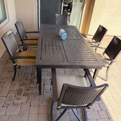 Sale Photo Thumbnail #249: Outdoor patio dining set featuring a dark metal slatted table and six woven-back armchairs with seat cushions.