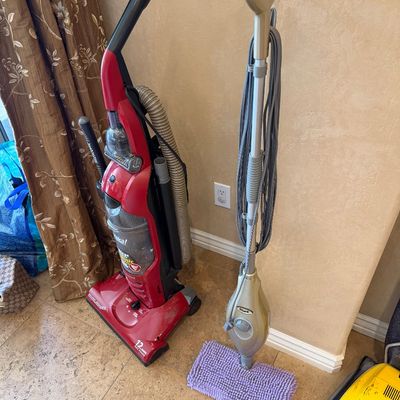 Sale Photo Thumbnail #152: Red Eureka upright vacuum cleaner standing next to a gold Shark steam mop on a tile floor.