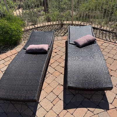Sale Photo Thumbnail #240: Pair of modern dark brown woven resin wicker lounge chairs, each topped with a small purple throw pillow on a brick patio.