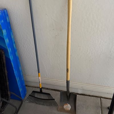 Sale Photo Thumbnail #66: Two long-handled yard tools leaning against a wall, including a metal leaf rake and an HDX square-point transfer shovel with a wooden handle.
