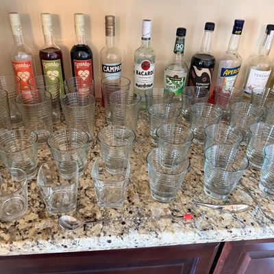 Sale Photo Thumbnail #32: Collection of barware including various clear ribbed glassware, bar tools, and open bottles of liquor lined up on a granite countertop.