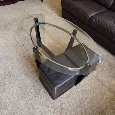 Sale Photo Thumbnail #226: Contemporary oval glass-top coffee table featuring a dark wood base with chrome accents and two nesting dark brown leather ottomans beneath.