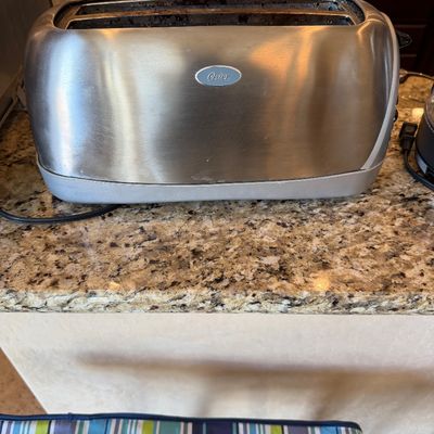 Sale Photo Thumbnail #189: Oster two-slot brushed stainless steel toaster sitting on a granite countertop, showing minor signs of previous use.