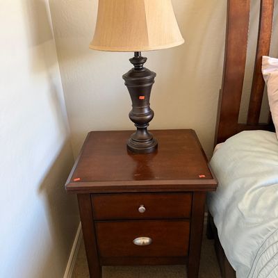 Sale Photo Thumbnail #115: Traditional dark wood two-drawer nightstand topped with a turned dark metal table lamp featuring a beige bell shade.