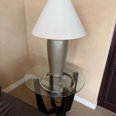 Sale Photo Thumbnail #231: Contemporary brushed metallic urn-form table lamp with white fabric shade, sitting atop a round glass-top accent table with dark wood legs.