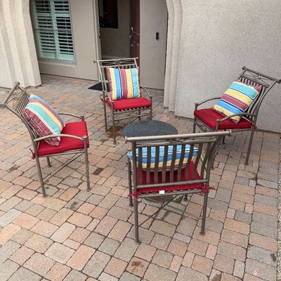Sale Photo Thumbnail #5: Four metal patio armchairs with red seat cushions and colorful striped throw pillows, arranged around a small round side table on a paved courtyard.