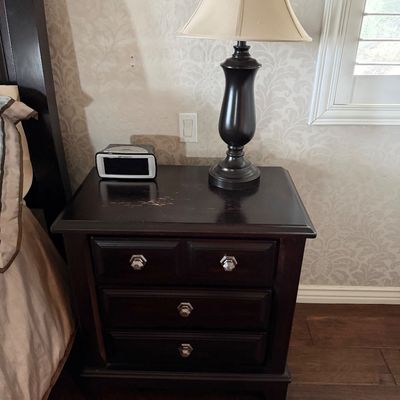 Sale Photo Thumbnail #20: Dark wood three-drawer nightstand with geometric metal pulls, featuring a traditional black urn-style table lamp and a digital alarm clock on top.