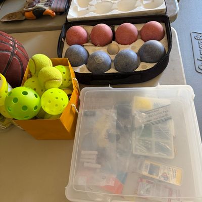 Sale Photo Thumbnail #94: Assortment of outdoor game equipment and garage supplies, including a bocce ball set in a case, tennis balls, pickleballs, work gloves, and a plastic organizer box with hardware.
