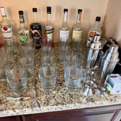 Sale Photo Thumbnail #33: Collection of home bar essentials including clear ribbed glassware, stainless steel cocktail shakers, jiggers, and assorted liquor bottles.