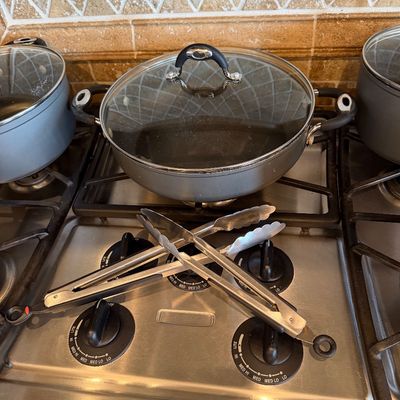 Sale Photo Thumbnail #183: Gray non-stick deep skillet with glass lid alongside additional cookware and two stainless steel kitchen tongs.