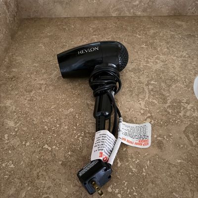 Sale Photo Thumbnail #125: Black Revlon handheld hair dryer with attached power cord and safety tags, resting on a bathroom vanity.