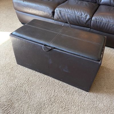 Sale Photo Thumbnail #232: Dark brown leather storage ottoman featuring contrast stitching and fabric pull tabs, set in front of a matching leather sofa.