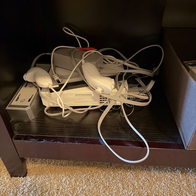 Sale Photo Thumbnail #60: White Nintendo Wii console with power brick, sensor bar, cabling, and two nunchuk controllers stored on an entertainment center shelf.