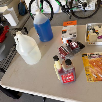 Sale Photo Thumbnail #95: Assorted household supplies including plastic jugs, Feit light bulbs, steel wool, Sta-Bil fuel stabilizer, and Sylvania decorative bulbs, displayed on a folding table in front of a green and black bicycle.