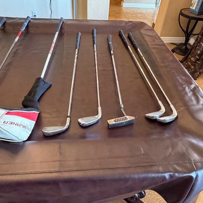 Sale Photo Thumbnail #255: Assortment of modern golf clubs including a TaylorMade AeroBurner driver with headcover, various irons, and a putter displayed on a brown table cover.