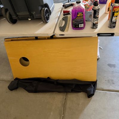 Sale Photo Thumbnail #108: Collection of garage and cleaning supplies on folding tables, including a rolling cooler, spray bottles, car wash liquid, and a set of wooden cornhole boards.