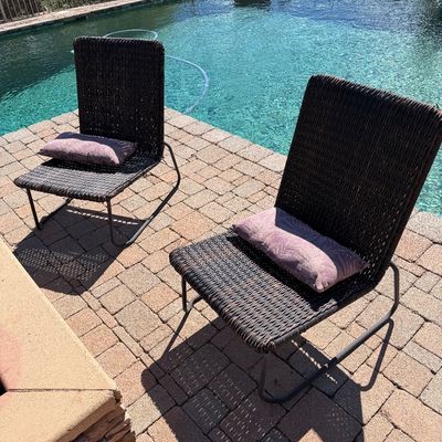 Sale Photo Thumbnail #236: Pair of contemporary outdoor woven lounge chairs with metal frames and lumbar accent pillows, situated poolside on a paved patio.