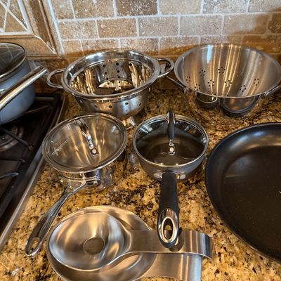 Sale Photo Thumbnail #185: Collection of stainless steel cookware and kitchen accessories including strainers, saucepans with glass lids, a frying pan, ladle, and spoon rest.