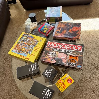 Sale Photo Thumbnail #53: Collection of board games and jigsaw puzzles on a glass coffee table, including Monopoly Cheaters Edition, Cards Against Humanity, and Smart Ass.