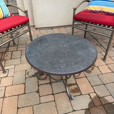 Sale Photo Thumbnail #4: Round metal patio side table with scrolled legs, flanked by two matching metal outdoor armchairs featuring red seat cushions and colorful striped pillows.