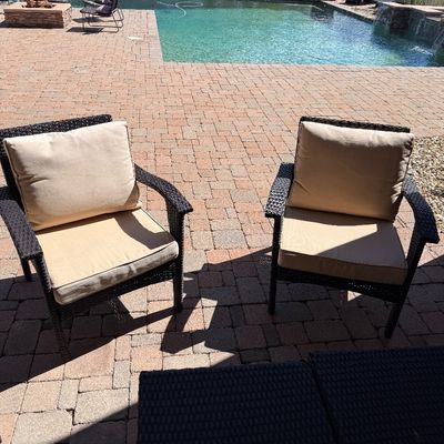 Sale Photo Thumbnail #243: Pair of dark brown resin wicker outdoor armchairs with beige seat and back cushions, situated on a paved patio.