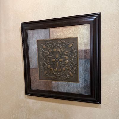 Sale Photo Thumbnail #118: Framed mixed-media wall art featuring a central embossed metal medallion with a raised floral pattern, set against a patchwork background of textured tiles and enclosed in a dark wooden frame.