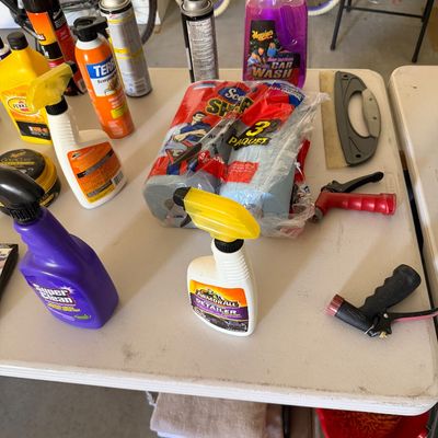 Sale Photo Thumbnail #98: Assortment of automotive care products and cleaning supplies, including Meguiar's car wash, Armor All detailer, Super Clean spray, shop towels, and hose nozzles arranged on a folding table.