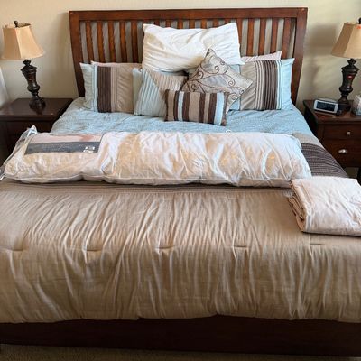 Sale Photo Thumbnail #114: Mission-style wooden bed frame with slatted headboard, dressed in neutral linens and decorative accent pillows, flanked by matching nightstands and table lamps.