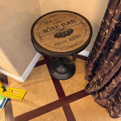 Sale Photo Thumbnail #31: Round rustic accent table featuring a wood-look top with black stenciled 'Welcome to Our Wine Bar' typography and a dark metal base.