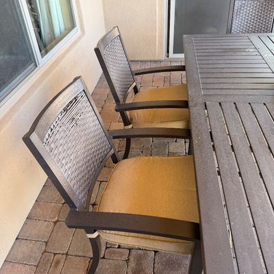 Sale Photo Thumbnail #242: Outdoor patio dining set featuring a slatted top table and dark metal armchairs with woven backs and tan seat cushions.