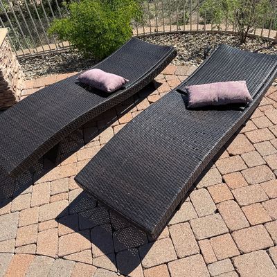 Sale Photo Thumbnail #237: Pair of dark brown woven synthetic wicker curved lounge chairs with accent pillows, situated outdoors on a paved patio.