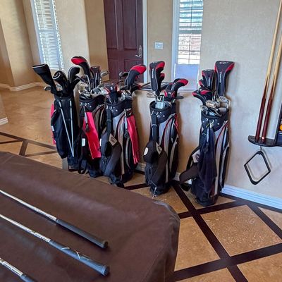 Sale Photo Thumbnail #257: Multiple sets of modern golf clubs in black and red stand bags lined up along a room wall.
