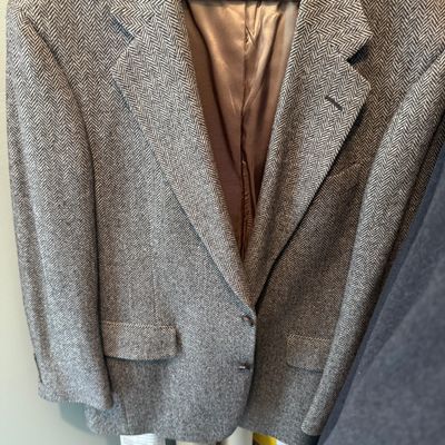 Sale Photo Thumbnail #91: Men's grey herringbone tweed blazer jacket hanging on a wooden coat hanger. Features notch lapels, flap pockets, and a full interior lining in good vintage condition.
